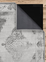 Vista de la alfombra 'Alma' mostrando la capa inferior antideslizante y el frente y reverso de la capa superior, que es lavable en lavadora. El diseño incluye un medallón central intrincado y detalles arabescos en tonos grises y plateados, con un efecto desgastado que le da un aire vintage.